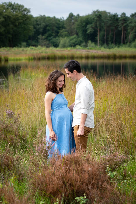 Een zwangere vrouw in een blauwe jurk tegenover haar partner voor een meertje tussen de heide. Zwangerschapsshoot.