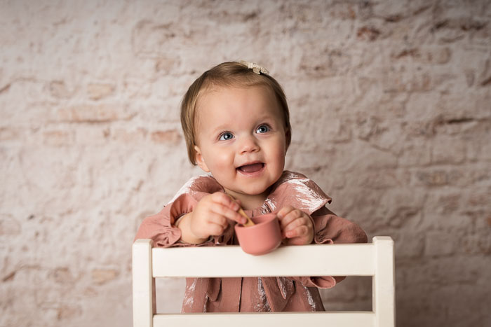 Meisje in roze jurk, staand achter een stoel voor een witte muur. Portretfoto.