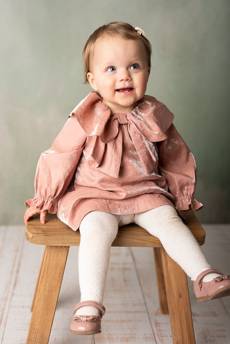 Portret foto van een meisje van 1 jaar oud in een roze jurkje, zittend op een krukje voor een groene achtergrond.