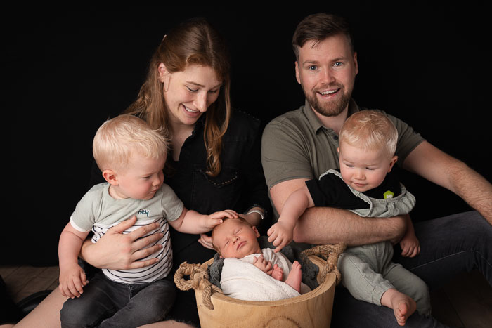 Portretfoto van een vader en moeder met een tweeling en een pasgeboren baby slapend in een mandje.