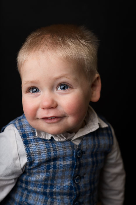 Portretfoto van een jongetje met een lieve blik, in een blauw giletje voor een zwarte achtergrond.