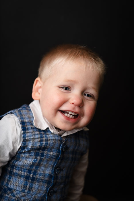 Portretfoto van een jongetje die lacht, in een blauw giletje voor een zwarte achtergrond.